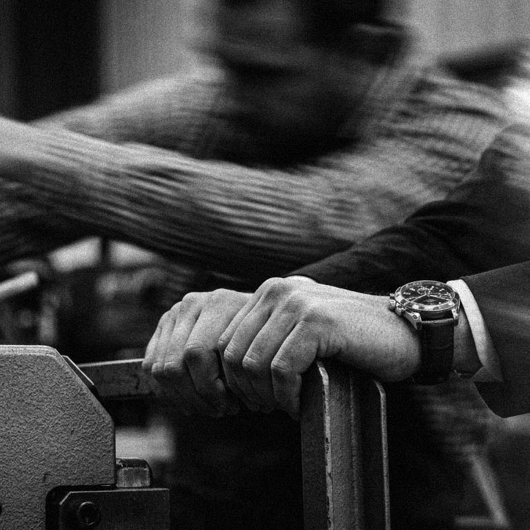 An owner's hand resting on a shop machine while somebody moves past behind, in black and white.