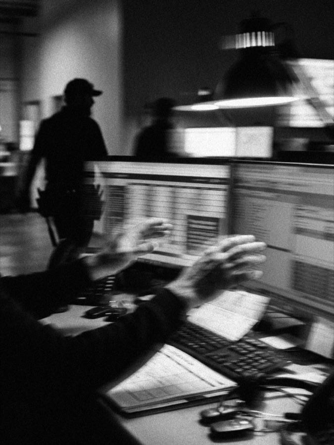 A dispatch desk at dusk, hands sorting schedule sheets beside a keyboard as a uniformed technician walks past in the background, in black and white.