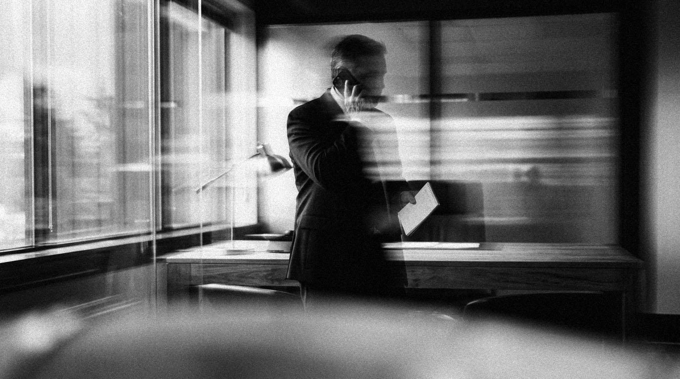 An owner on a phone call near an office window, a notepad in the other hand, in black and white.