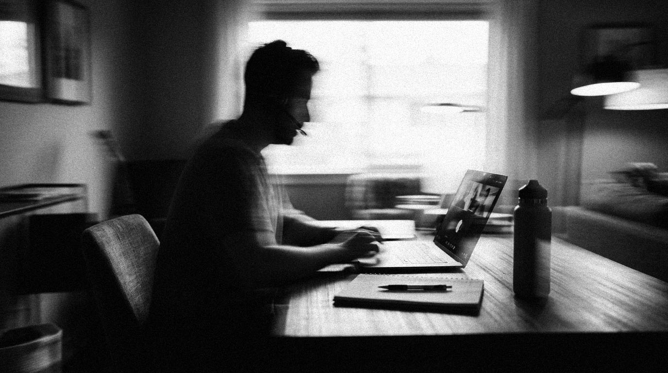A remote worker with a headset on a video call at a home-office desk, a notepad and water bottle beside the laptop, in black and white.