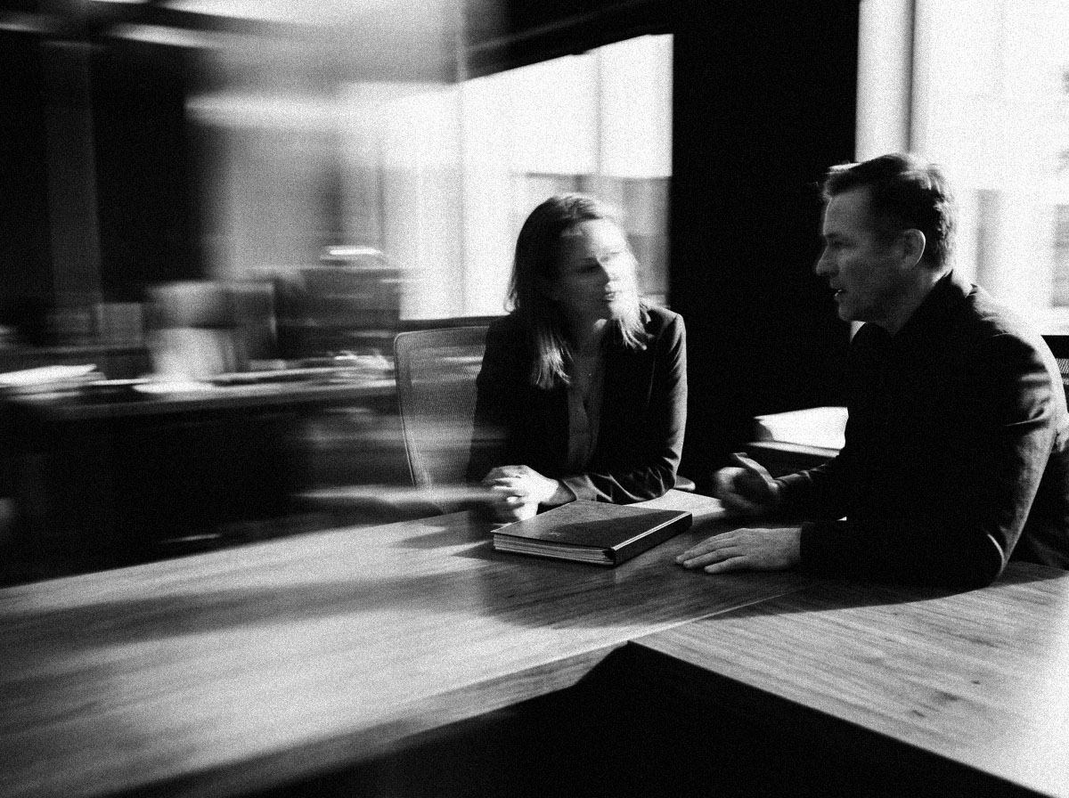 Two people talking across the corner of a desk with a closed report between them, in black and white.