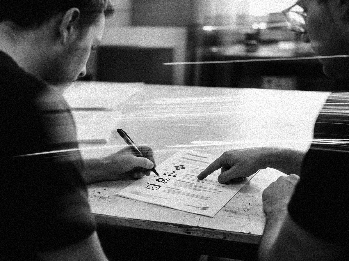 Two people leaning over a printed process sheet on a workbench, one marking it with a pen while the other points at a step, in black and white.