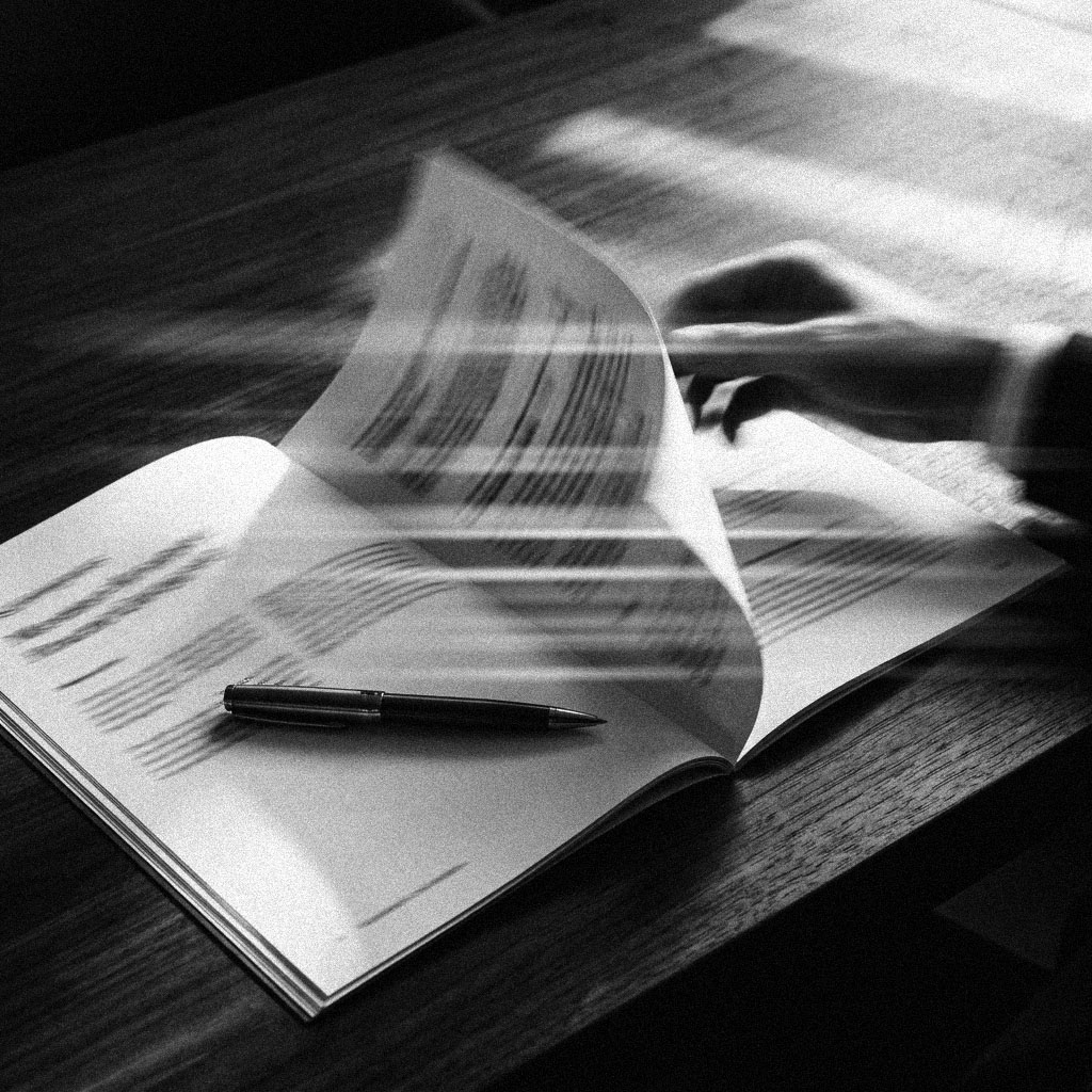 A hand turning a page of a printed report open on a dark desk, a pen resting across the page underneath, in black and white.