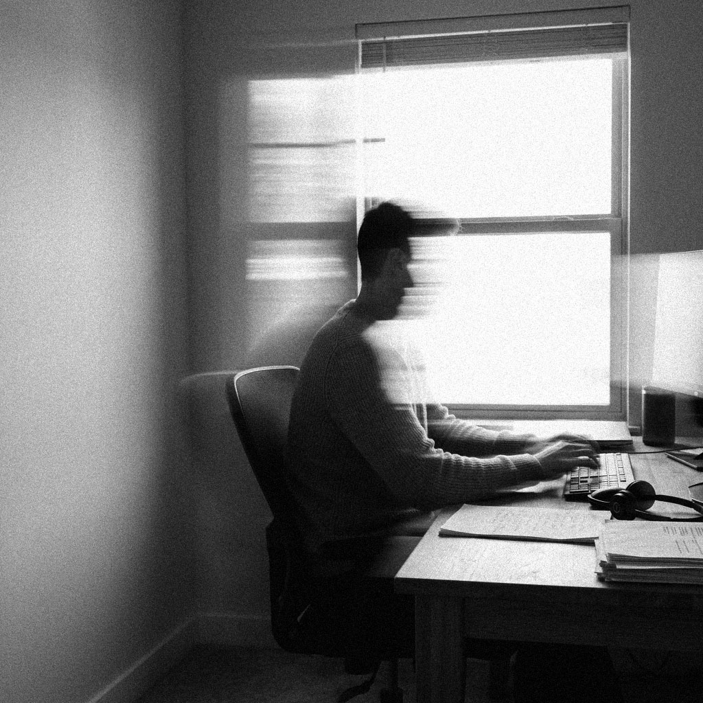 A remote worker typing at a spare-room desk with a headset set down beside a stack of processed paperwork, in black and white.